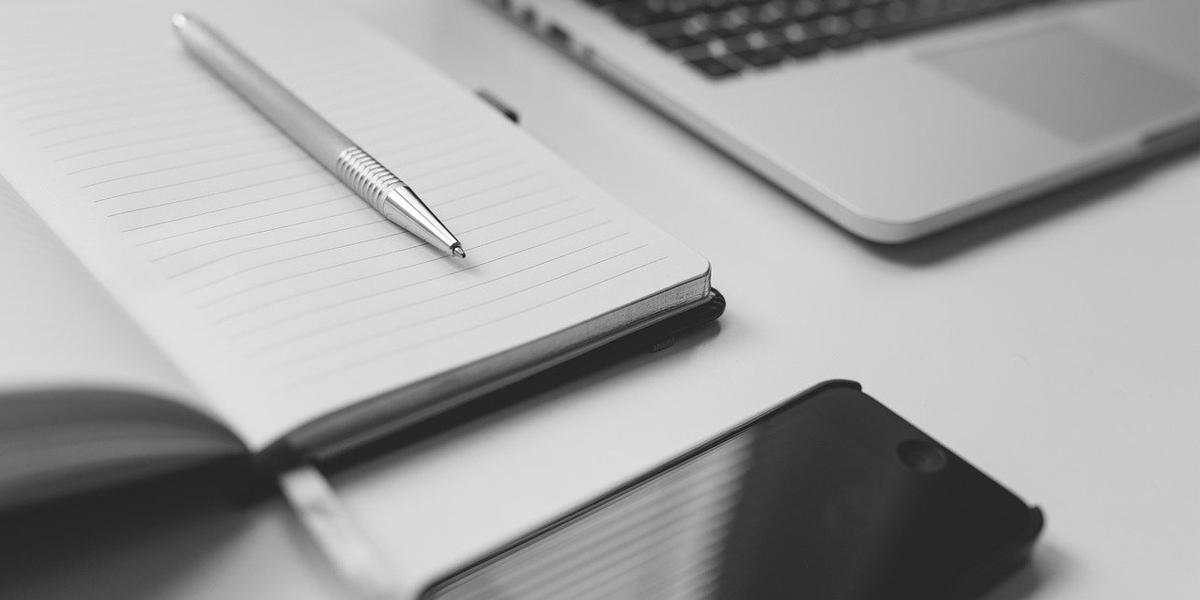 Black and white image of an open notebook next to a laptop and smartphone.